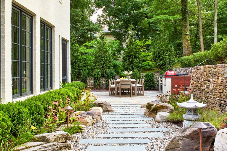 The integration of classical garden elements with a looser, but no less specific approach addresses the husband and wife’s different design sensibilities. The entry garden pairs formal shapes with low plantings and perennial flowers. At the rear, traditional dry stack retaining walls surrounding the entertaining area appear beautifully contemporary in their expression. Two patios—one for cooking and dining, the other for repose around a fireplace—are connected via a walkway of pavers surrounded by river stone, which in turn are bordered by traditional boxwood, boulders, ornamental grasses, and perennial flowers. To reinforce the inside/outside connection, an axial path of stone in running bond leads from the front door through the house to culminate in the back garden. Upon arriving at the terrace stair, the stone path gives way to an irregular pattern that defines the patio areas.