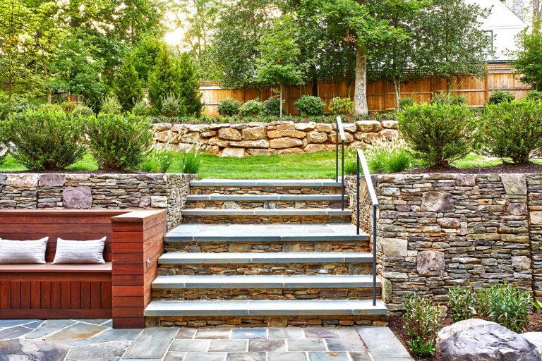 The integration of classical garden elements with a looser, but no less specific approach addresses the husband and wife’s different design sensibilities. The entry garden pairs formal shapes with low plantings and perennial flowers. At the rear, traditional dry stack retaining walls surrounding the entertaining area appear beautifully contemporary in their expression. Two patios—one for cooking and dining, the other for repose around a fireplace—are connected via a walkway of pavers surrounded by river stone, which in turn are bordered by traditional boxwood, boulders, ornamental grasses, and perennial flowers. To reinforce the inside/outside connection, an axial path of stone in running bond leads from the front door through the house to culminate in the back garden. Upon arriving at the terrace stair, the stone path gives way to an irregular pattern that defines the patio areas.