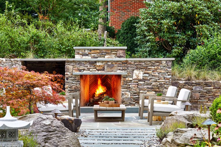 The integration of classical garden elements with a looser, but no less specific approach addresses the husband and wife’s different design sensibilities. The entry garden pairs formal shapes with low plantings and perennial flowers. At the rear, traditional dry stack retaining walls surrounding the entertaining area appear beautifully contemporary in their expression. Two patios—one for cooking and dining, the other for repose around a fireplace—are connected via a walkway of pavers surrounded by river stone, which in turn are bordered by traditional boxwood, boulders, ornamental grasses, and perennial flowers. To reinforce the inside/outside connection, an axial path of stone in running bond leads from the front door through the house to culminate in the back garden. Upon arriving at the terrace stair, the stone path gives way to an irregular pattern that defines the patio areas.