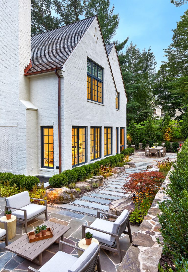 The integration of classical garden elements with a looser, but no less specific approach addresses the husband and wife’s different design sensibilities. The entry garden pairs formal shapes with low plantings and perennial flowers. At the rear, traditional dry stack retaining walls surrounding the entertaining area appear beautifully contemporary in their expression. Two patios—one for cooking and dining, the other for repose around a fireplace—are connected via a walkway of pavers surrounded by river stone, which in turn are bordered by traditional boxwood, boulders, ornamental grasses, and perennial flowers. To reinforce the inside/outside connection, an axial path of stone in running bond leads from the front door through the house to culminate in the back garden. Upon arriving at the terrace stair, the stone path gives way to an irregular pattern that defines the patio areas.