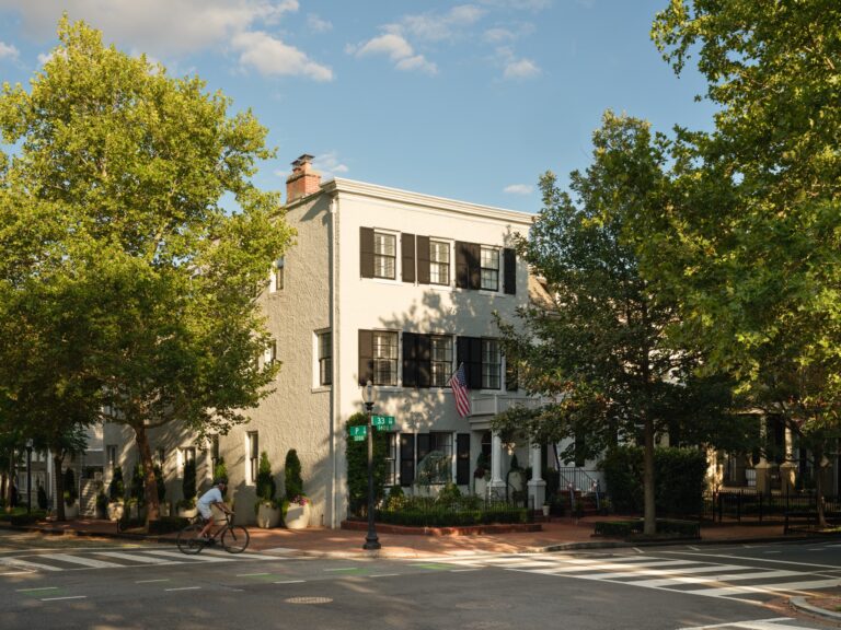 A white three-story house on a street corner, with black shutters and an American flag hanging from the porch. A person on a bicycle rides through the intersection in the foreground.