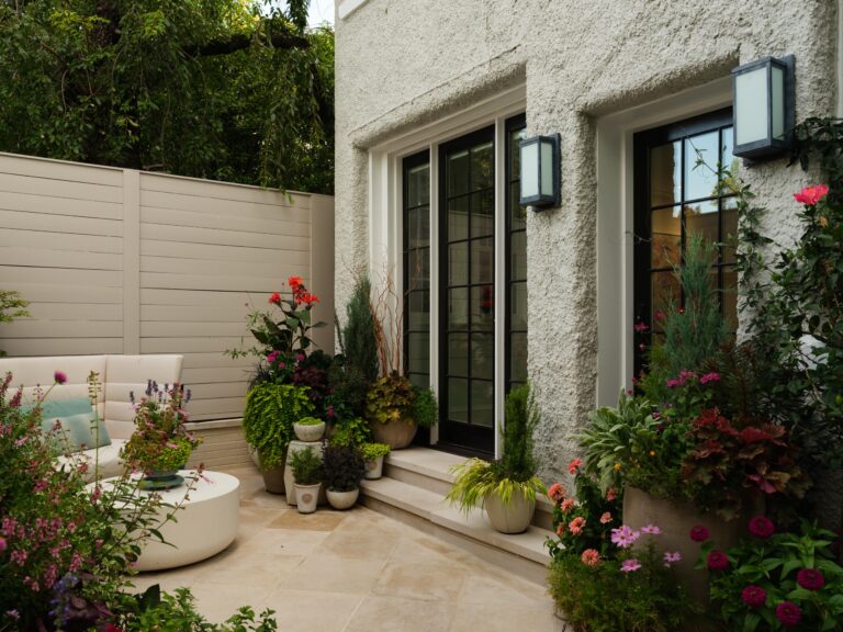 A patio area next to a white house with large glass doors and black frames. A beige sofa sits on a stone floor near a collection of potted plants and flowers.
