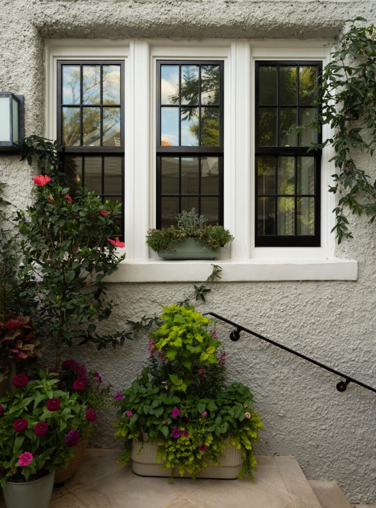 A triple window with black panes and white trim is set in a textured stucco wall. Below the window, two large planters overflow with a variety of colorful flowers and greenery, and a black metal handrail extends along the stone steps.