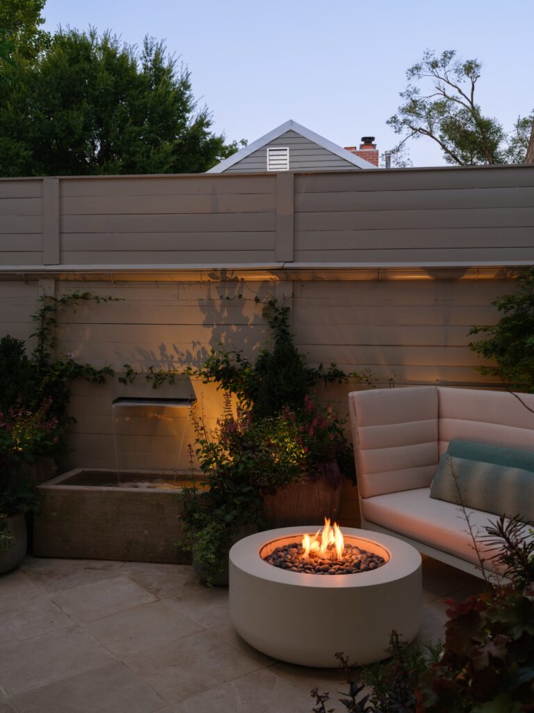 A circular stone fire pit with a glowing flame sits on a patio next to a curved outdoor sofa. Behind them, a stone water feature spills into a basin against a tall wooden privacy fence adorned with climbing plants.