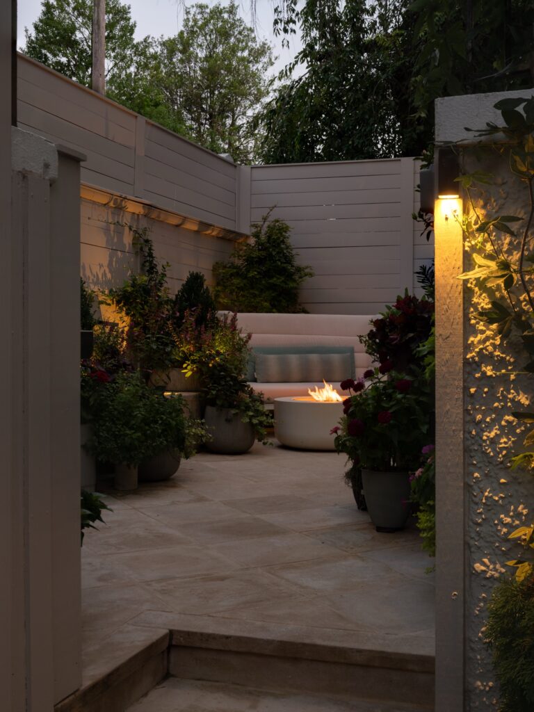 A patio nook features a circular fire pit in front of a curved bench seating area. The space is surrounded by potted plants and enclosed by light-colored horizontal fencing, illuminated by warm outdoor wall lights.