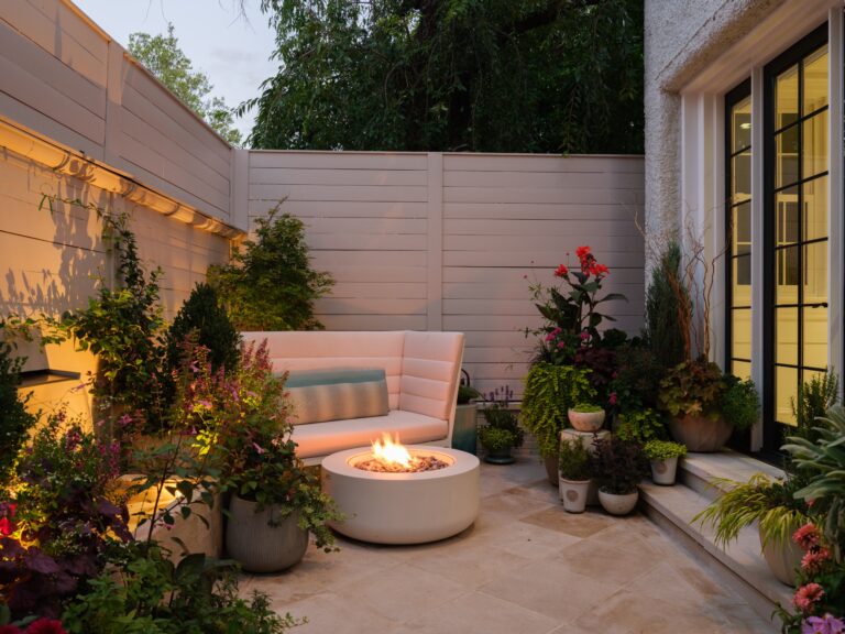 A patio seating area features a white curved outdoor sofa and a round fire pit. The space is surrounded by a light-colored privacy fence and various potted plants against the wall of a home.