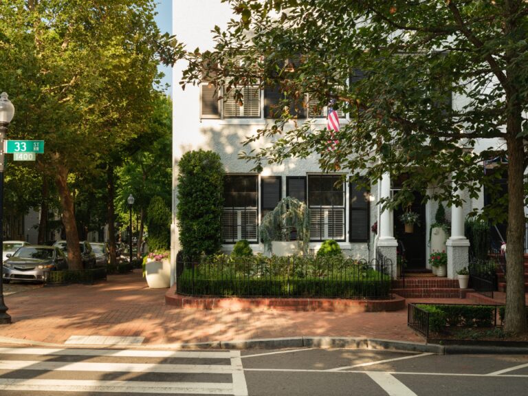 A white townhome with black shutters and a small front garden surrounded by a metal fence. A street sign in the foreground marks the corner of 33rd Street NW.