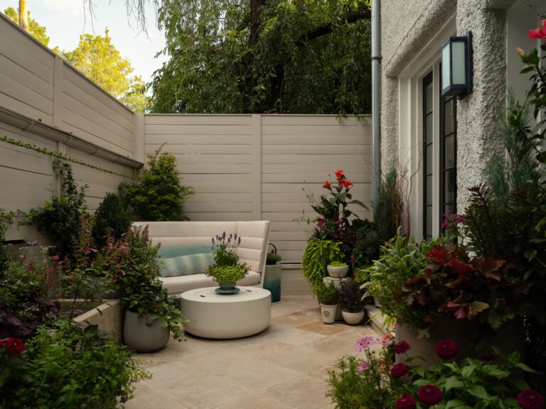 A cozy patio seating area featuring a curved white sofa and a matching circular coffee table. The space is enclosed by light-colored wooden fencing and lush container gardens filled with various flowers and greenery.