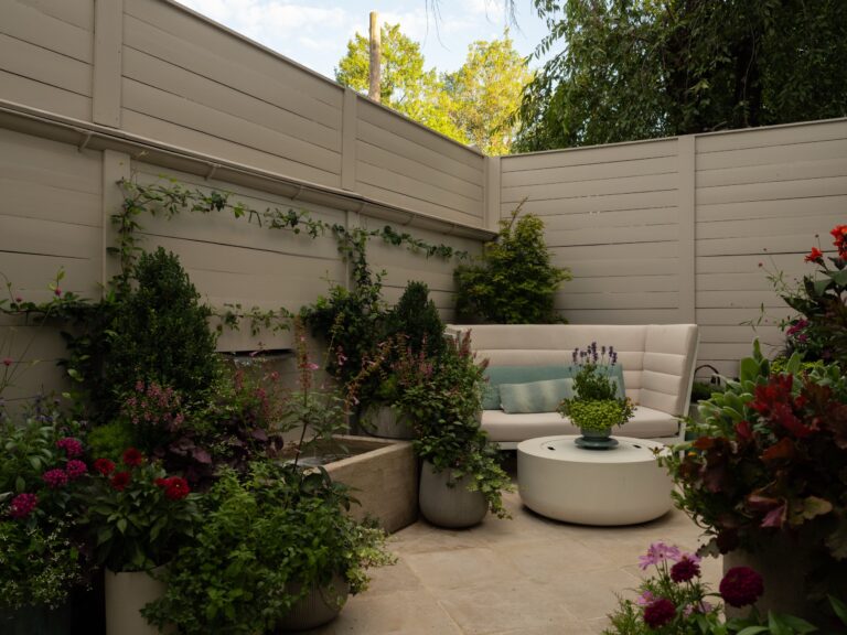 A modern outdoor seating area features a curved beige sofa and a round white coffee table on a stone patio. The space is enclosed by horizontal wood fencing and surrounded by lush potted plants and a stone water feature.