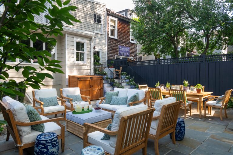 A patio with light wood furniture including chairs with white cushions and green patterned pillows arranged around a central ottoman. A dining table set for four sits in the background near a black fence, with a banner reading Dont Give Up The Ship hanging on the adjacent house.