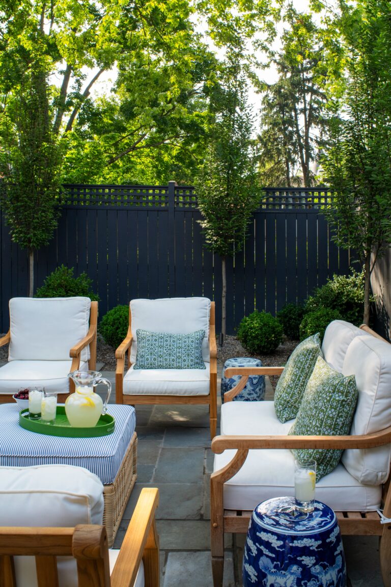 Outdoor patio seating with white cushions and patterned green pillows arranged around a blue-and-white striped ottoman. A green tray with a pitcher and glasses of lemonade sits on the ottoman, set against a dark fence and lush trees.