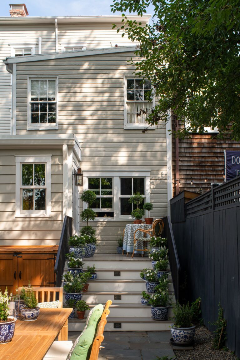 The back exterior of a light-colored wood-paneled house featuring a short staircase decorated with several potted plants. At the top of the stairs sits a small table and chair, while a larger dining table and chairs occupy the foreground patio.
