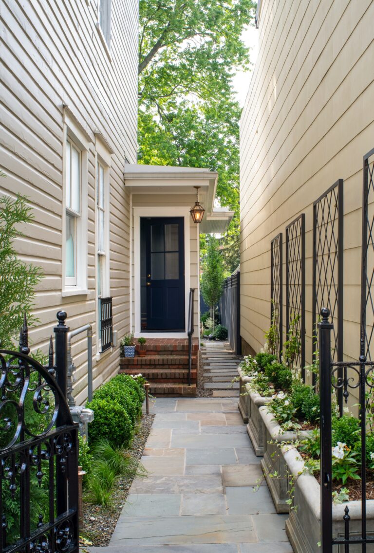 A narrow stone walkway leads to a dark blue front door with a small porch and lantern. Flanking the path are metal garden trellises, stone planters with greenery, and an open wrought-iron gate.