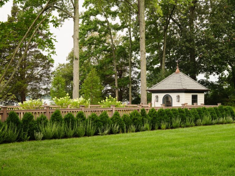 A small, light-colored garden house with a hip roof sits behind a wooden fence. The property is surrounded by a line of manicured hedges and tall trees, with a large green lawn in the foreground.