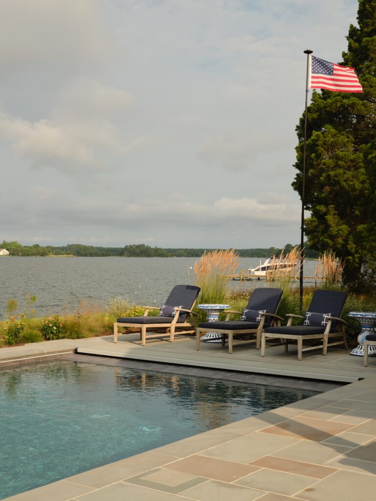 Three lounge chairs sit on a poolside patio overlooking a calm body of water. An American flag on a tall pole flies to the right, near a boat docked in the distance.