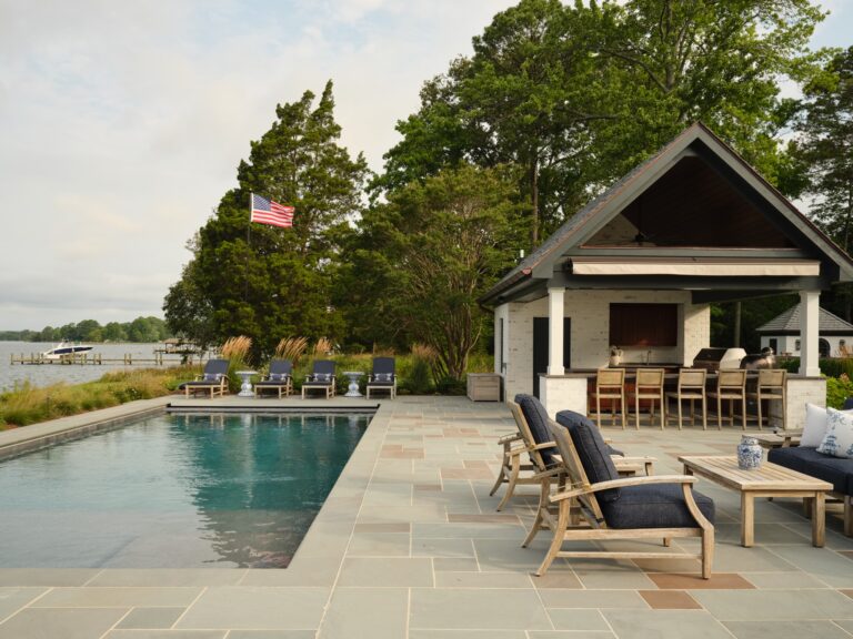 A rectangular swimming pool sits on a stone patio next to an outdoor kitchen and covered seating area. Several lounge chairs face the water, where an American flag flies near the shoreline.