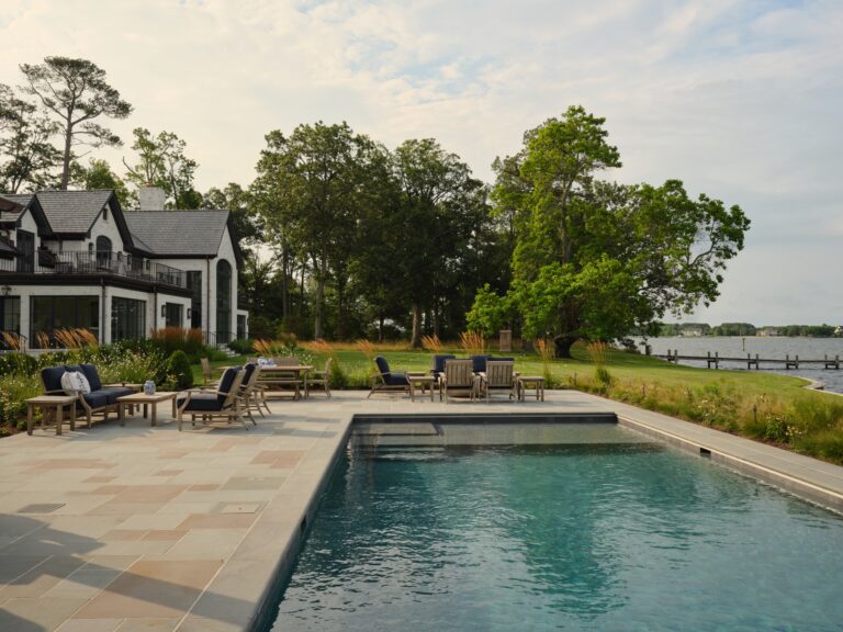 A backyard swimming pool area with outdoor lounge furniture on a stone patio. A multi-story white house stands to the left, with large trees and a calm body of water in the background.