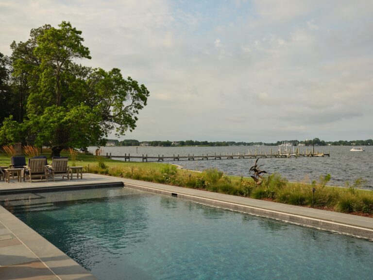 An infinity pool overlooking a wide river with a long wooden pier extending into the water. Patio chairs sit on a stone deck beside the pool, and a metal sculpture stands in a garden bed nearby.