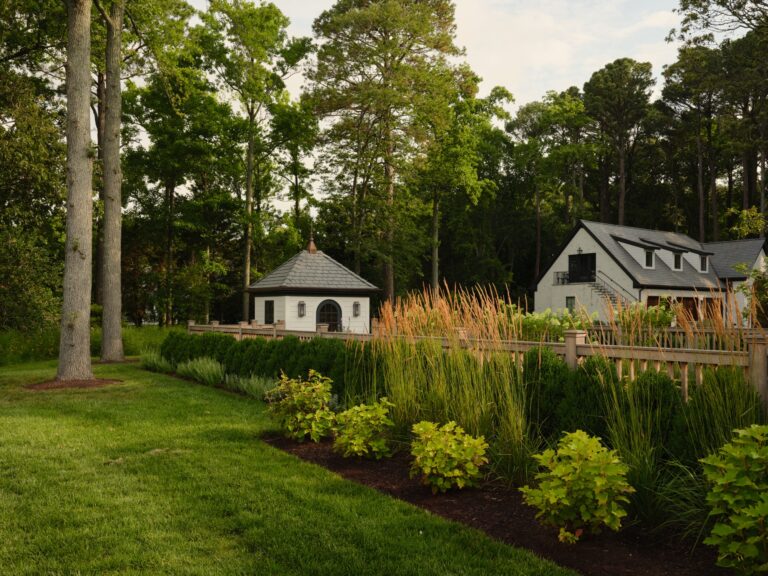 A white gazebo with a dark roof stands in a lush garden next to a wood fence and ornamental grasses. A white two-story house with a dormer roof is visible in the background among tall trees.