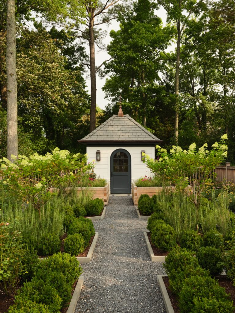 A small, white garden shed with a gray roof sits at the end of a gravel path. The path is lined with symmetrical boxwood shrubs and raised garden beds containing flowering plants, all set against a backdrop of tall green trees.