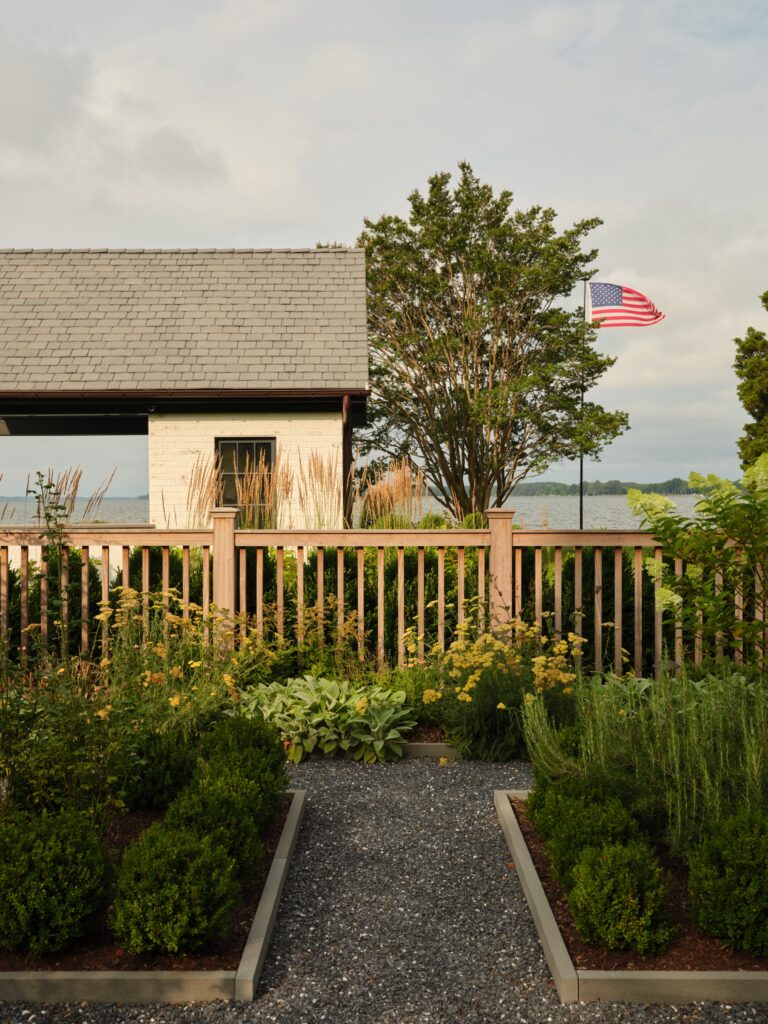 A gravel path leads through a garden toward a wooden fence and a small white building by the water. An American flag flies on a pole next to a tree behind the fence.