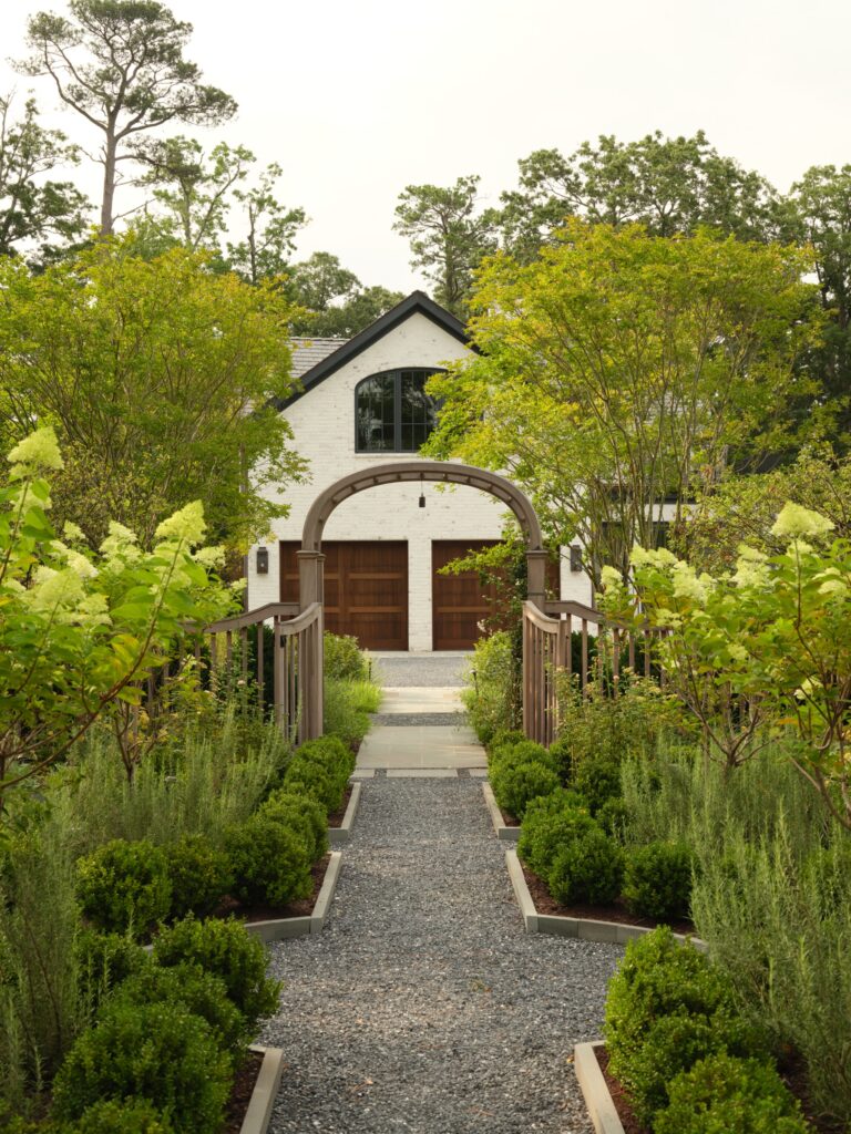 A gravel path leads through a garden with manicured shrubs toward a white brick carriage house. A wooden arch frames the entrance to the path, which is lined by green trees and flowering bushes.