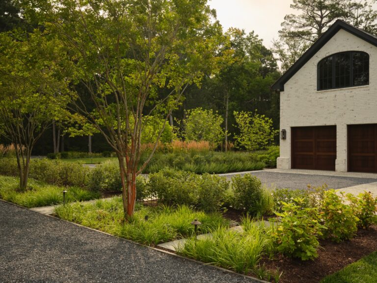 A white brick garage with dark arched windows and wooden doors sits next to a landscaped garden. A gravel driveway leads to the garage, framed by small trees, manicured shrubs, and low-voltage path lights.