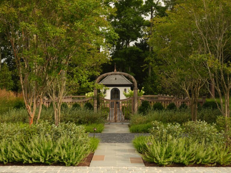A stone path leads to a wooden arched gate opening into a garden with a small white building at the center. The garden is enclosed by a low wooden fence and surrounded by lush green trees and shrubs.