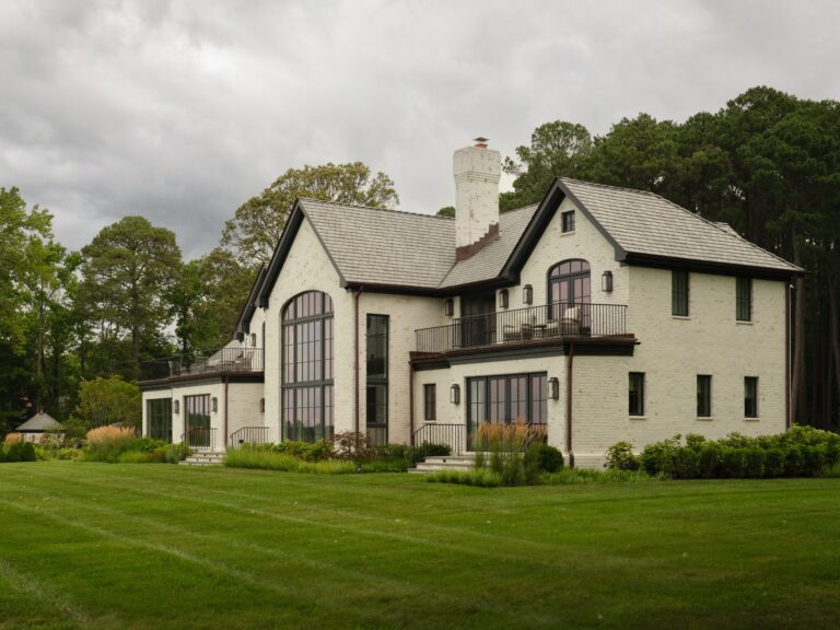 A large, white brick house with a dark roof and black-framed windows sits on a grassy lawn. The home features a prominent two-story arched window, a second-story balcony with railings, and a tall brick chimney.