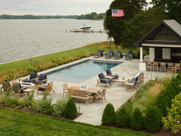A swimming pool sits on a patio next to an outdoor bar and a large body of water. The deck features multiple seating areas with wood chairs, tables, and blue cushions, with an American flag flying nearby.