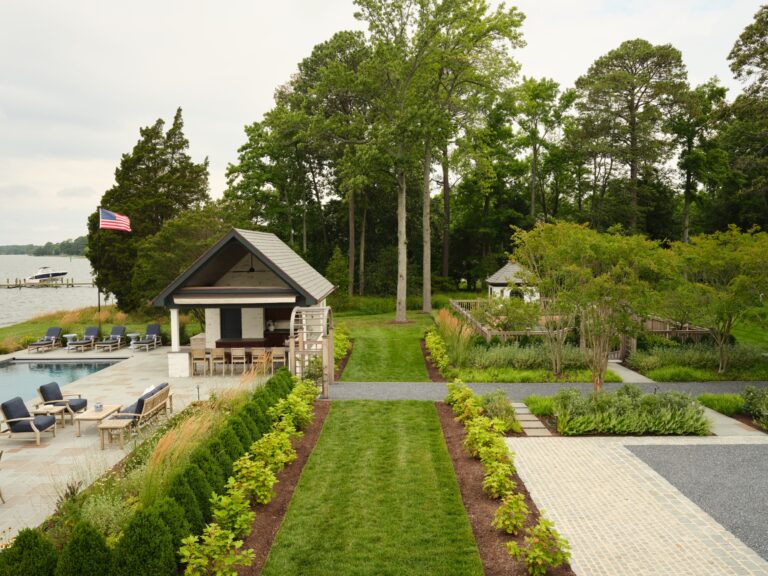 A waterfront backyard features a stone patio with a pool and an outdoor kitchen pavilion near the water. A central lawn path separates the pool area from a paved garden space surrounded by mature trees and lush landscaping.