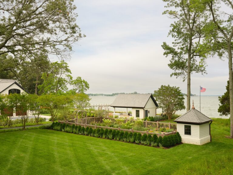 A fenced garden sits between a small pool house and a stone cottage near a large lake. A detached garage is on the left, and an American flag flies near the water.