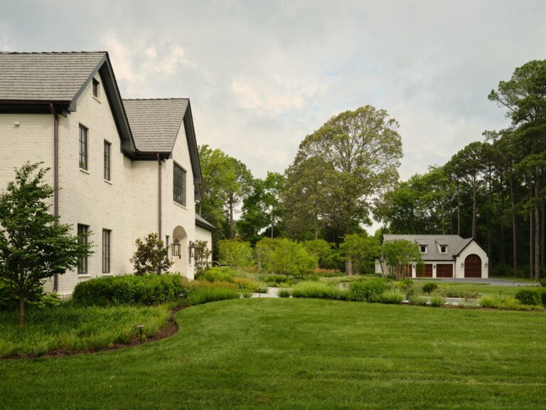 A white brick house with a dark gabled roof sits next to a manicured lawn and garden. In the background, a detached garage with two arched wooden doors is nestled among tall trees.