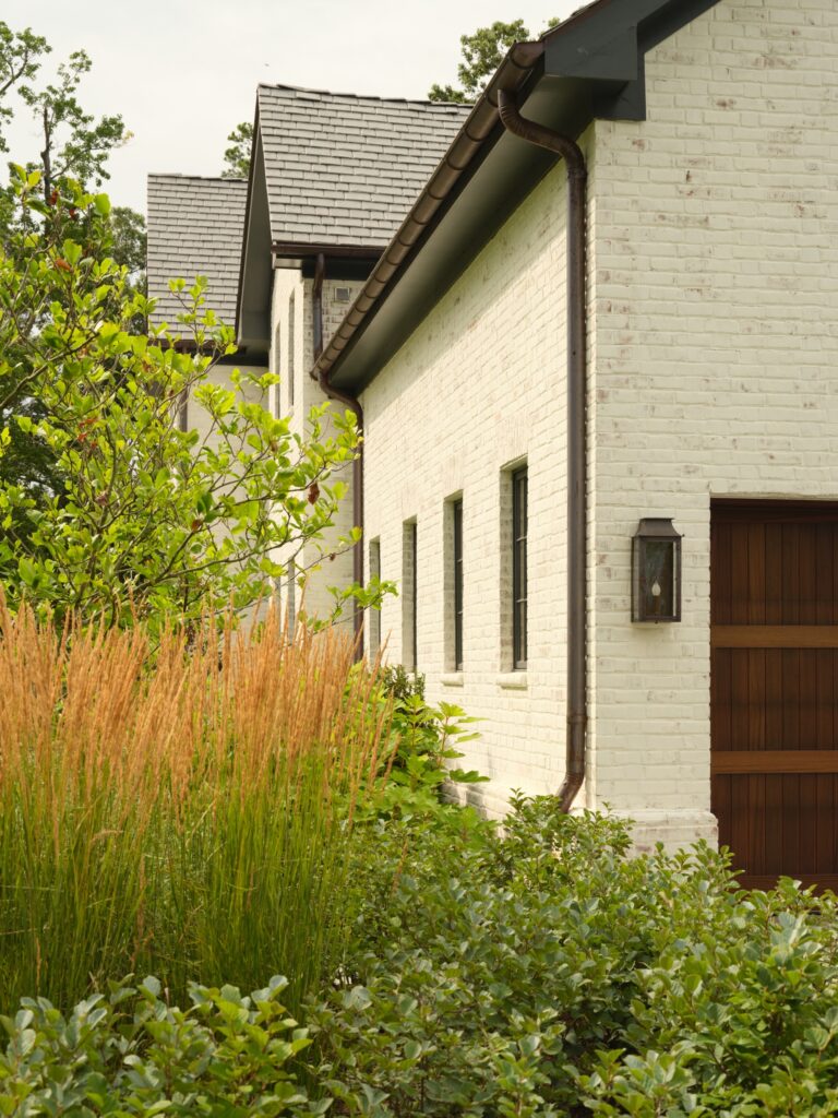 A modern home with white painted brick siding and a dark brown wooden garage door. Tall ornamental grasses and shrubs grow in the foreground, with dark gutters running along the roofline.