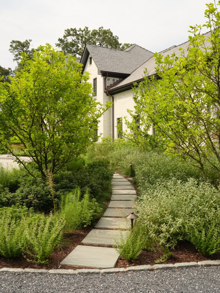 A stone stepping-stone path leads through a lush garden to a white brick house. The path is bordered by green shrubs, small trees, and two landscape lights.