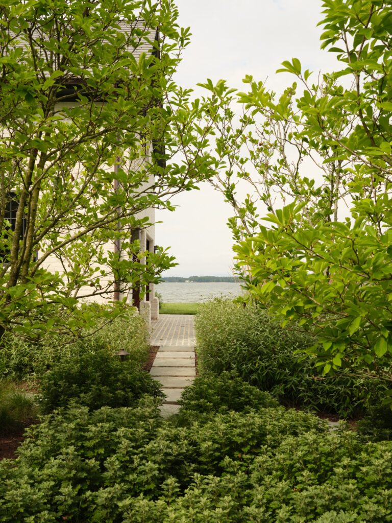 A stone path leads between green trees and bushes toward a lake. The path passes the side of a building and opens onto a view of the water.