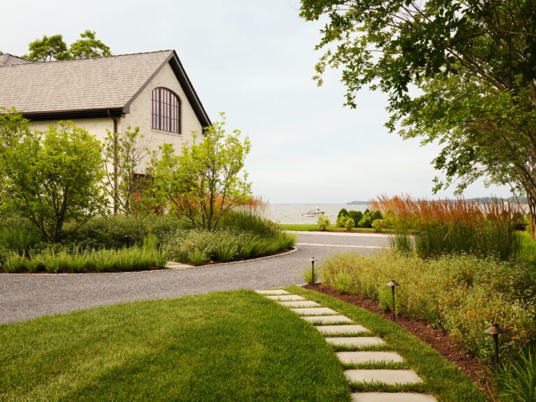 A white brick building with a dark shingled roof stands beside a gravel driveway. A stone path leads through a green lawn toward the water, where a small boat is visible in the distance.