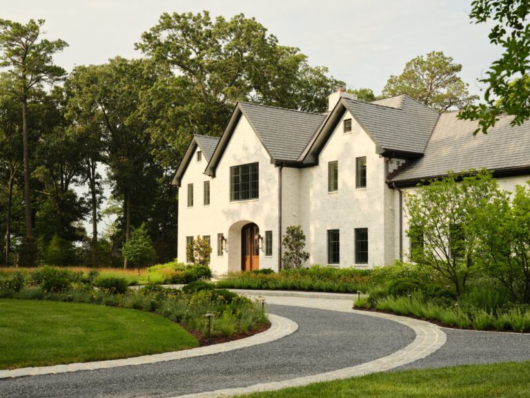 A two-story white brick house with a dark shingled roof and a wooden front door. A curved gravel driveway leads to the entrance, surrounded by landscaped gardens and a lawn in front of a dense treeline.